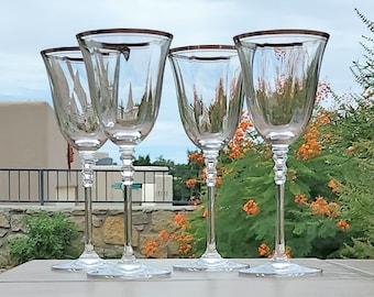 May include: Four clear glass wine glasses with a gold rim. The glasses are sitting on a light brown table.