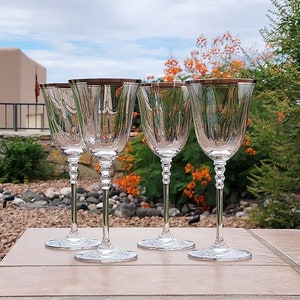 May include: Set of four clear glass wine glasses with a gold rim. The glasses are arranged in a row on a light brown surface.