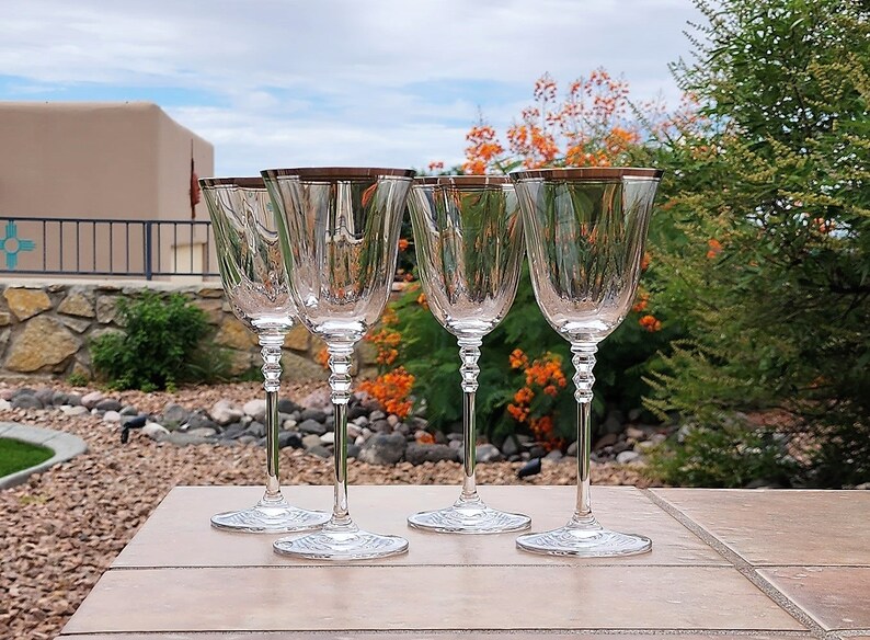 May include: Set of four clear glass wine glasses with a gold rim. The glasses are arranged in a row on a light brown surface.