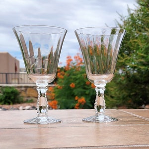 May include: Two clear glass wine glasses with beaded stems. The glasses are sitting on a tiled surface.