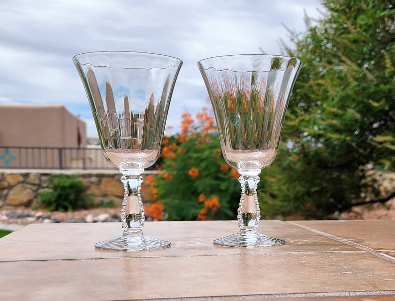 May include: Two clear glass wine glasses with beaded stems. The glasses are sitting on a tiled surface.