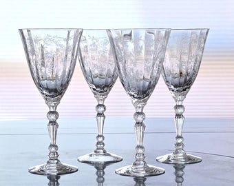 May include: Four clear crystal wine glasses with etched floral designs. Each glass features a round base, a decorative stem with a ball detail, and a flared bowl. The glasses are arranged in a row, reflecting light.