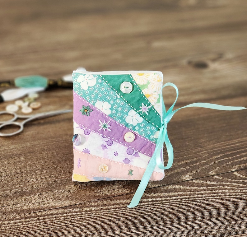 Puede incluir: Un peque&ntilde;o coj&iacute;n para alfileres de tela acolchada con una cinta azul claro. El coj&iacute;n est&aacute; hecho de tela rosa, morada y verde con lunares blancos y patrones florales. Tiene varios botones cosidos en la parte delantera.