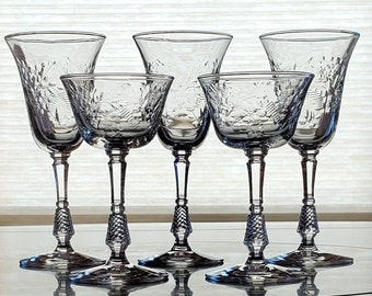 May include: Five clear glass stemmed drinking glasses with intricate etched designs. The glasses are arranged in a row on a table.