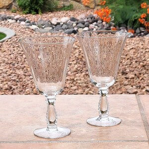 May include: Two clear glass wine glasses with a textured, ribbed design and beaded stems. The glasses are sitting on a brown tiled surface.