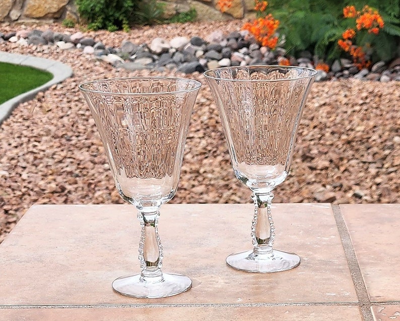 May include: Two clear glass wine glasses with a textured, ribbed design and beaded stems. The glasses are sitting on a brown tiled surface.