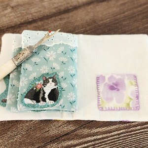 Puede incluir: Un estuche para agujas de tela blanca con estampado floral azul y dise&ntilde;o de gato blanco y negro. El estuche tiene un peque&ntilde;o parche floral morado y un enhebrador de agujas de hueso blanco.