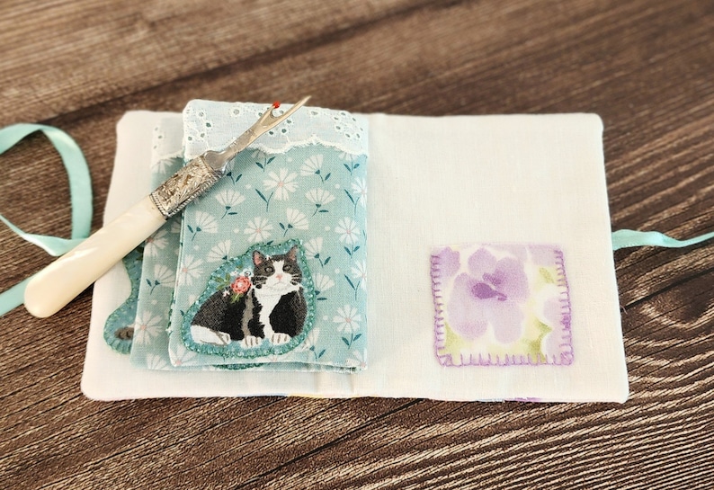 Puede incluir: Un estuche para agujas de tela blanca con estampado floral azul y dise&ntilde;o de gato blanco y negro. El estuche tiene un peque&ntilde;o parche floral morado y un enhebrador de agujas de hueso blanco.