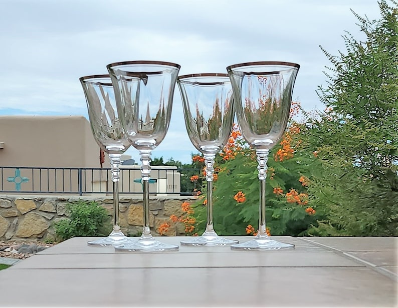 May include: Four clear glass wine glasses with a gold rim. The glasses are sitting on a light brown table.