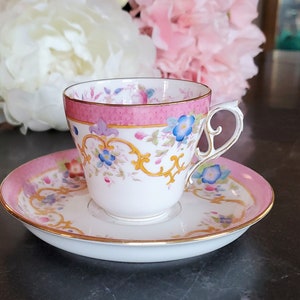 May include: A pink and white teacup and saucer set with a floral pattern and gold accents. The cup has a delicate handle and the saucer has a scalloped edge.
