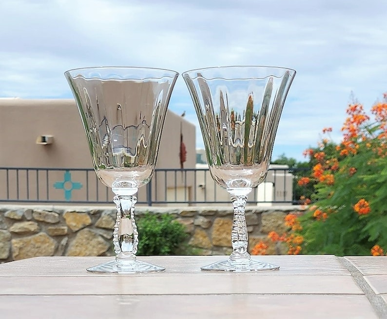 May include: Two clear glass wine glasses with a fluted design and a beaded stem. The glasses are sitting on a wooden table.
