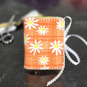 May include: A small, handmade fabric book with an orange plaid pattern and white daisy flowers. The edges are stitched with yellow thread. A white ribbon is attached to the book. The book is approximately 2 inches tall.