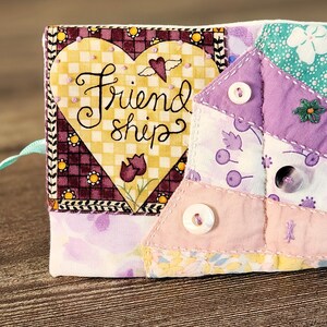 Puede incluir: Una bolsa de coraz&oacute;n de tela acolchada con un dise&ntilde;o cosido de un coraz&oacute;n y las palabras "Friendship". La bolsa est&aacute; hecha de varias telas estampadas en tonos de morado, rosa y verde. Tiene un lazo de cinta y tres botones blancos.