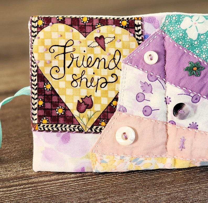 Puede incluir: Una bolsa de coraz&oacute;n de tela acolchada con un dise&ntilde;o cosido de un coraz&oacute;n y las palabras "Friendship". La bolsa est&aacute; hecha de varias telas estampadas en tonos de morado, rosa y verde. Tiene un lazo de cinta y tres botones blancos.