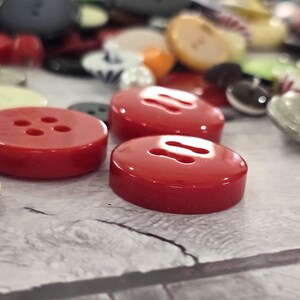 以下が含まれることがあります： さまざまな色とサイズのボタンのコレクション。4つの穴がある明るい赤色のボタンがいくつかあり、黒、クリーム、シルバーのボタンと一緒に焦点が当てられています。ボタンは、テクスチャ加工された明るい色の表面に散らばっています。