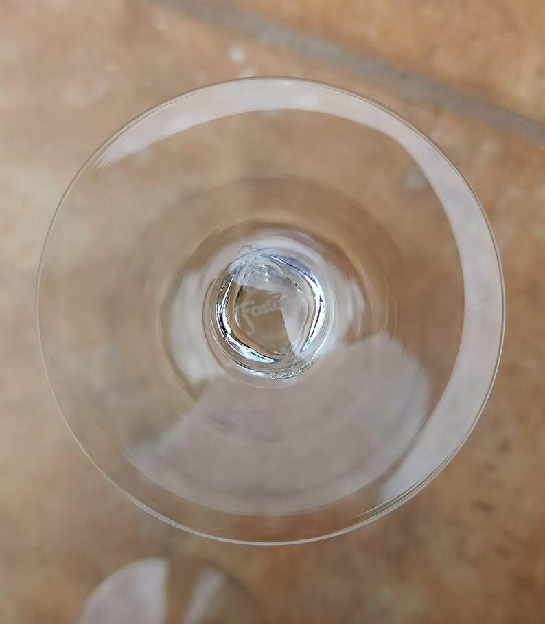 May include: The underside of a clear glass wine glass with a small, round, clear glass base. The base has the word "Fosta" etched into it.