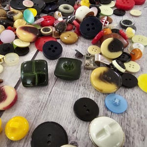 以下が含まれることがあります： さまざまな形、サイズ、色のボタンのコレクション。ボタンには、黒、白、黄色、赤、緑があります。4つの穴があるボタンもあれば、2つまたは穴がないボタンもあります。ボタンは明るい木製の表面に散らばっています。