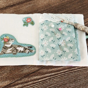 Puede incluir: Un libro de agujas de tela blanca con un dise&ntilde;o floral y una aplicaci&oacute;n de gato. El libro est&aacute; abierto para revelar una tela verde con un patr&oacute;n floral y varios alfileres. Tambi&eacute;n se puede ver un enhebrador de agujas plateado y blanco.
