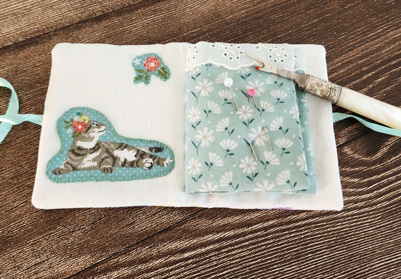 Puede incluir: Un libro de agujas de tela blanca con un dise&ntilde;o floral y una aplicaci&oacute;n de gato. El libro est&aacute; abierto para revelar una tela verde con un patr&oacute;n floral y varios alfileres. Tambi&eacute;n se puede ver un enhebrador de agujas plateado y blanco.