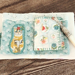 Puede incluir: Un peque&ntilde;o libro de tela blanco con un patr&oacute;n floral azul y blanco en el interior. El libro tiene un gato con una corona de flores en un lado y un patr&oacute;n floral en el otro. Una peque&ntilde;a herramienta de aguja de plata con mango blanco est&aacute; en el lado derecho del libro.
