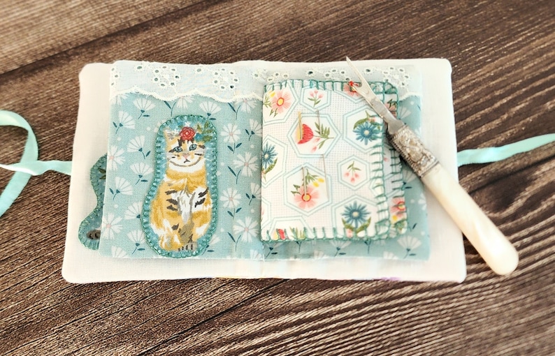 Puede incluir: Un peque&ntilde;o libro de tela blanco con un patr&oacute;n floral azul y blanco en el interior. El libro tiene un gato con una corona de flores en un lado y un patr&oacute;n floral en el otro. Una peque&ntilde;a herramienta de aguja de plata con mango blanco est&aacute; en el lado derecho del libro.