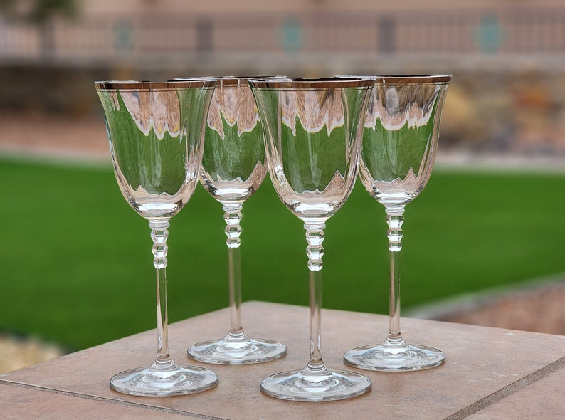 May include: Four clear glass wine glasses with silver rims sit on a brown surface. The glasses are empty and have a stemmed base.
