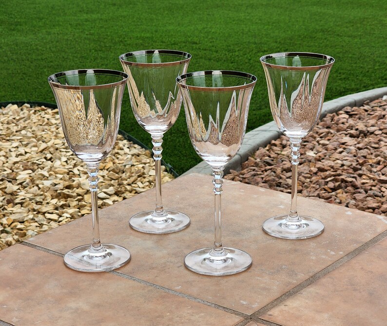 May include: Four clear glass wine glasses with silver rims. The glasses are arranged in a row on a brown tiled surface.