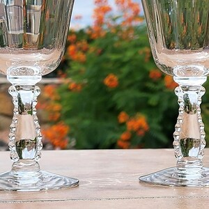 May include: Two clear glass stemmed goblets with beaded stems. The goblets are sitting on a light brown surface.