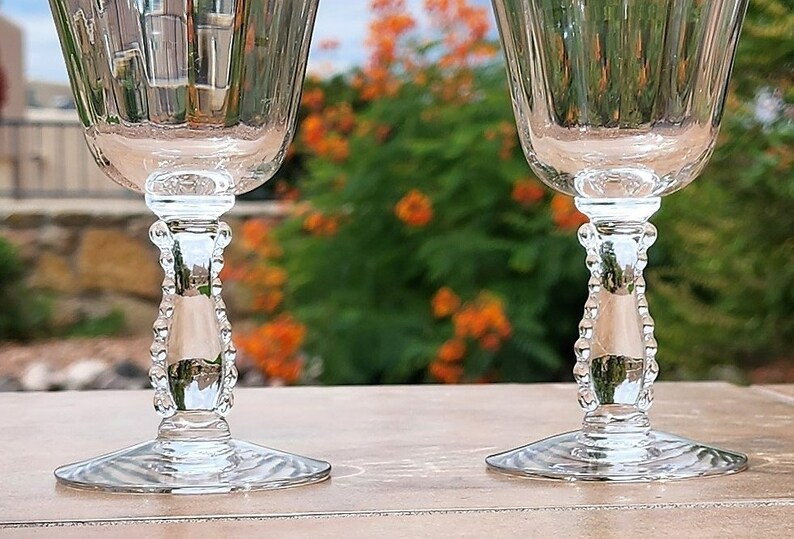 May include: Two clear glass stemmed goblets with beaded stems. The goblets are sitting on a light brown surface.