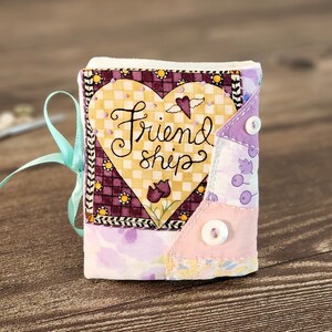 Puede incluir: Un peque&ntilde;o libro de tela hecho a mano con un parche en forma de coraz&oacute;n que dice "Friendship". El libro est&aacute; hecho de tela de patchwork en tonos de morado, rosa y amarillo.