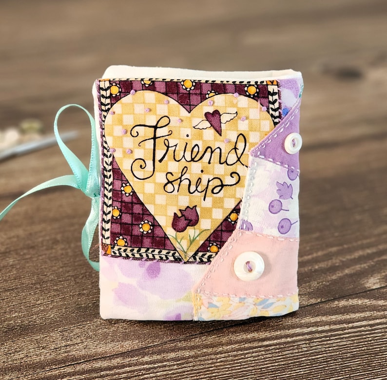 Puede incluir: Un peque&ntilde;o libro de tela hecho a mano con un parche en forma de coraz&oacute;n que dice "Friendship". El libro est&aacute; hecho de tela de patchwork en tonos de morado, rosa y amarillo.