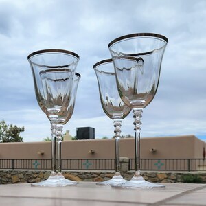 May include: Four clear glass wine glasses with a silver rim and a textured design. The glasses are arranged in a row on a table.
