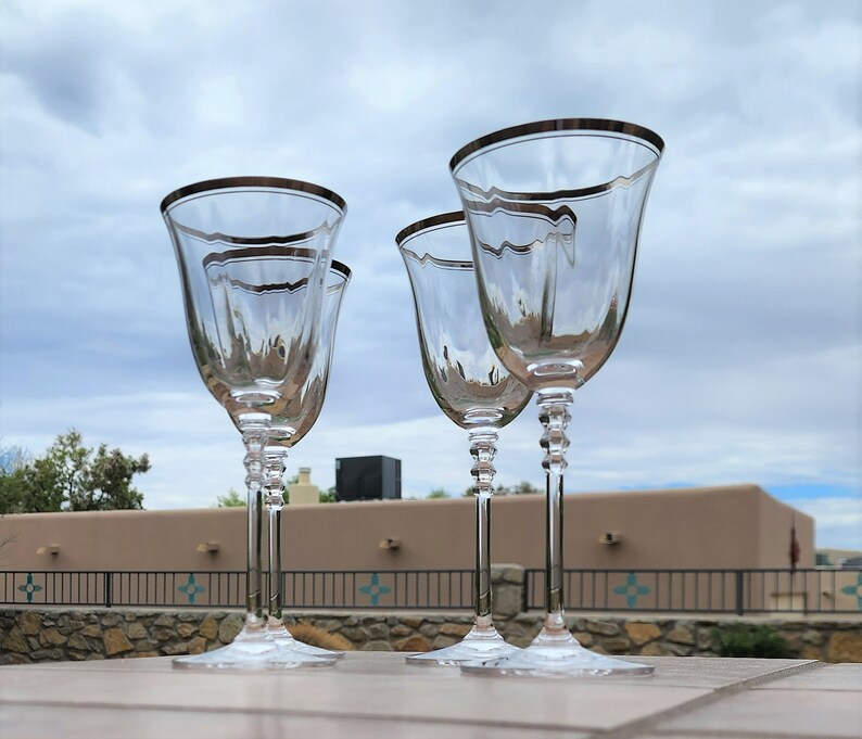 May include: Four clear glass wine glasses with a silver rim and a textured design. The glasses are arranged in a row on a table.