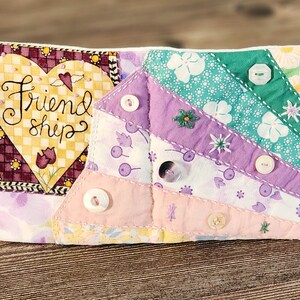 Puede incluir: Un libro de tela acolchado con un parche en forma de coraz&oacute;n que dice "Friendship". El libro est&aacute; decorado con botones y tela floral en tonos de rosa, morado y verde.