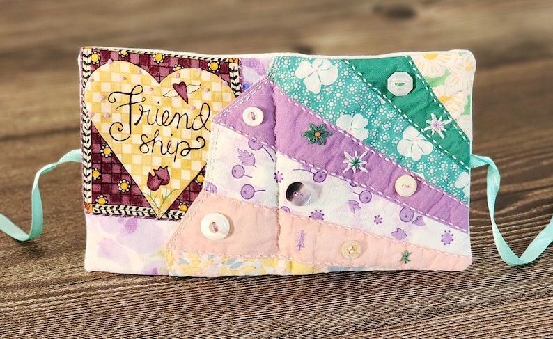 Puede incluir: Un libro de tela acolchado con un parche en forma de coraz&oacute;n que dice "Friendship". El libro est&aacute; decorado con botones y tela floral en tonos de rosa, morado y verde.