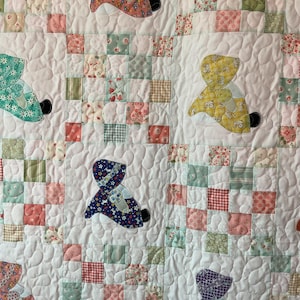 May include: A handmade quilt with a white background and colorful patchwork squares. The quilt features appliqued teddy bear figures in various patterned fabrics, including floral and gingham designs. The bears have black feet and are spaced across the quilt.