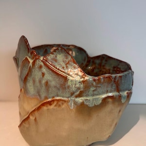 May include: A handmade ceramic bowl with a unique, organic shape. The bowl features a light tan base, transitioning to a turquoise and brown rim. The rim has a textured, uneven edge, adding to its artistic appeal.