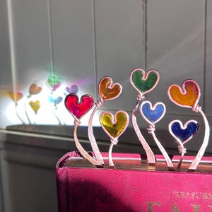 May include: A set of seven colorful glass heart-shaped bookmarks attached to wire stems. The bookmarks are arranged in a rainbow pattern, with red, orange, yellow, green, blue, indigo, and violet hearts.
