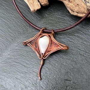 May include: A copper wire necklace with a white shell pendant shaped like a stingray. The necklace is on a brown leather cord.