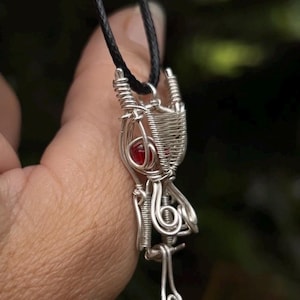 Peut inclure: Un pendentif en fil d'argent avec une pierre précieuse rouge, suspendu à un cordon noir.