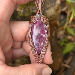 May include: A pendant featuring a marquise-shaped magenta and white stone encased in intricate copper wire wrapping. A small, round, iridescent stone is set at the top, just below the decorative wire bail, creating a detailed metalwork design.