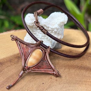 May include: A copper wire wrapped pendant featuring a light brown shell in the shape of a manta ray. The pendant is attached to a brown leather cord.
