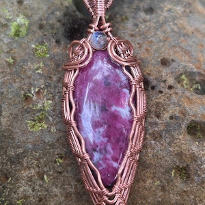 Peut inclure: Un pendentif composé d'une pierre magenta et blanche en forme de goutte, entourée d'un fil de cuivre finement tressé. Une petite pierre de lune ronde et irisée est fixée au sommet, intégrée au design détaillé de la bélière en cuivre.