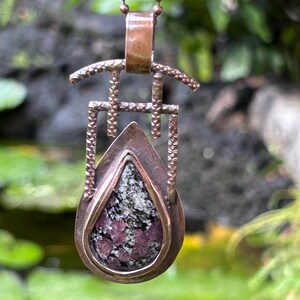 Peut inclure: Un pendentif en cuivre avec une pierre rose et grise en forme de larme sertie dans un cadre géométrique. Le pendentif est suspendu à une chaîne en cuivre.