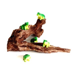 May include: Four small, green and yellow frog figurines are positioned on and around a piece of textured, brown driftwood. Three of the frogs are perched on the wood, while one frog sits separately in front of the branch on a white surface.