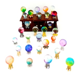 May include: A collection of colorful crystal balls on decorative stands. The spheres display a variety of colors, including red, blue, green, and orange. Some are displayed on a dark wooden cabinet, while others are arranged on a white surface.