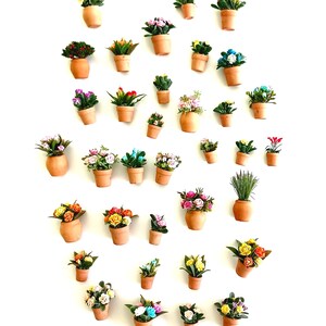May include: A collection of miniature terracotta pots, each containing a variety of small artificial flowers and greenery in diverse colors including pink, yellow, blue, red, and white, arranged in a scattered pattern against a plain white background.