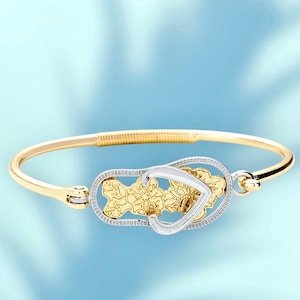 May include: Gold-tone bangle bracelet with a decorative centerpiece. The centerpiece features a gold floral design within a silver-tone heart-shaped frame. The bracelet has a simple, elegant design.