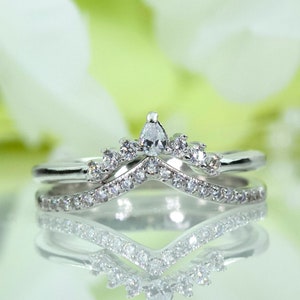 May include: A silver ring set with a delicate crown design featuring a pear-shaped center stone and smaller round stones. The ring is reflected in a mirror-like surface.