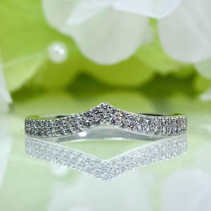 May include: A silver-toned ring with a chevron design, encrusted with small, sparkling stones. The ring's shape is mirrored in its reflection, creating a symmetrical image. The background is a soft, blurred mix of white and green.